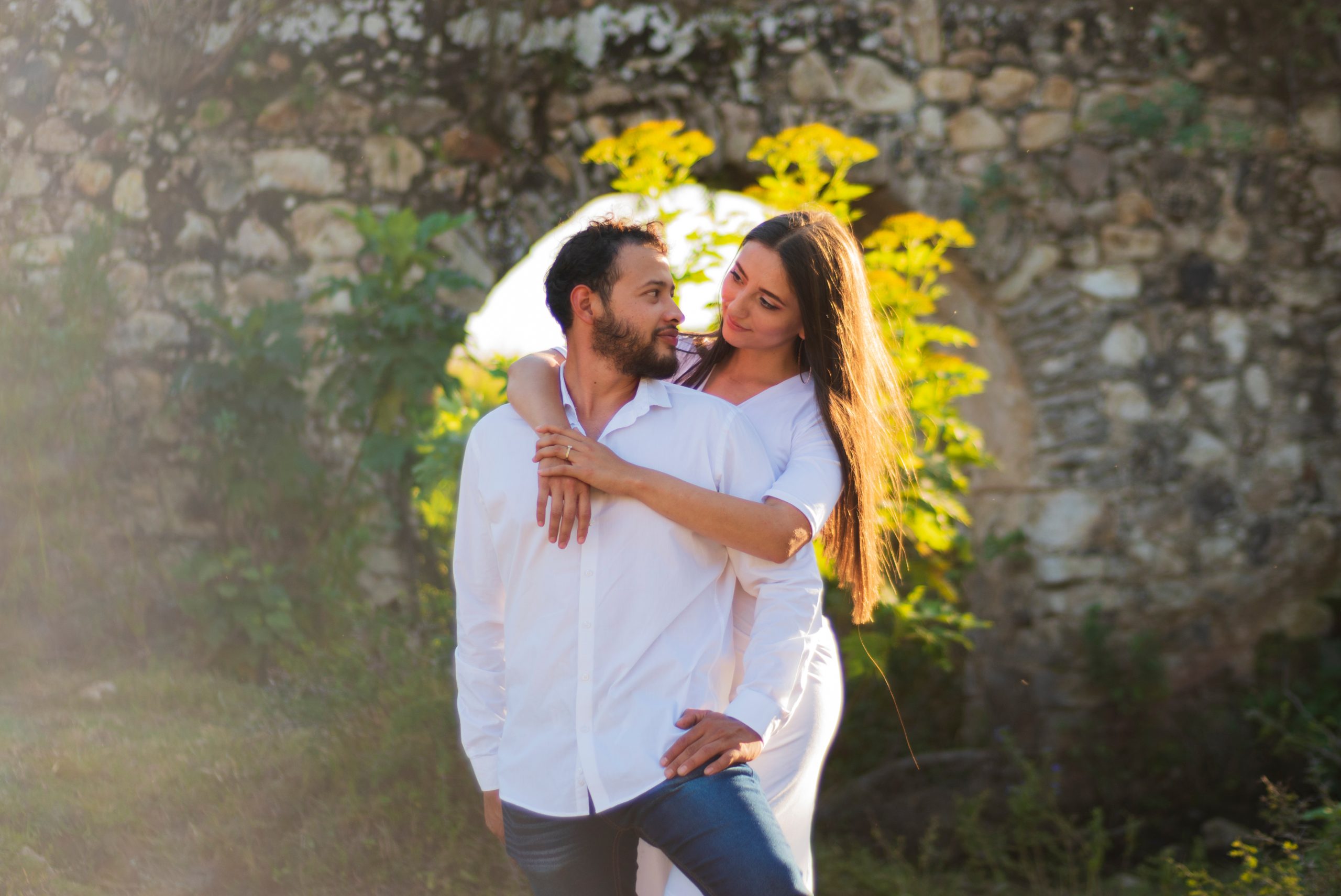 A romantic and lovely latin couple dressed in white with the ruins of a stone bridge behind and a beautiful sunset in the background